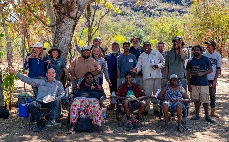 A large group of people waving at the camera. They are on Country. The group is a mixture of men and women, and some people are wearing ranger uniforms.